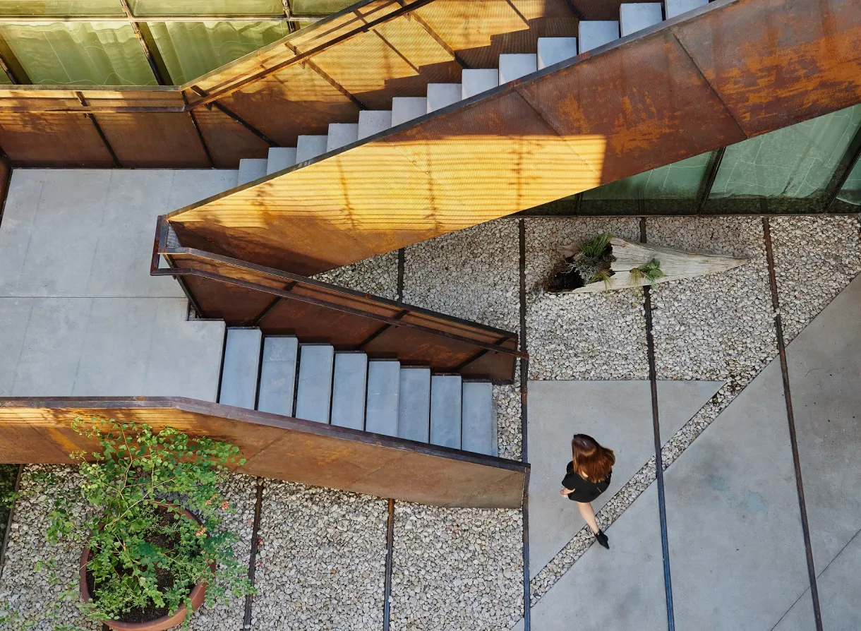 Overhead view of woman walking through minimalist courtyard with rusted metal stairs and stone paths